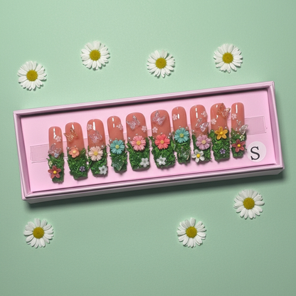 Set of decorative nail art with floral designs in a pink box on a light green background with daisies.