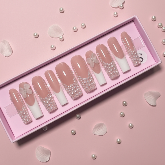 Set of decorative nail art tools in a pink box on a pink background with scattered pearls and petals.